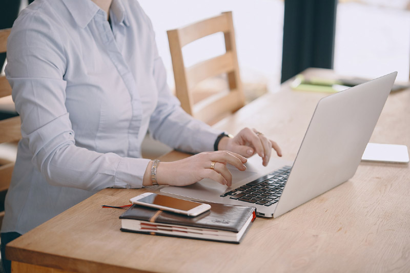 Person tippt auf Laptop an einem Holztisch mit Smartphone und Notizbüchern daneben