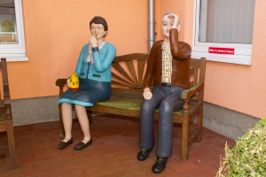 Zwei lebensgroße Figuren eines Mannes und einer Frau sitzen auf einer Holzbank vor einem orangefarbenen Haus