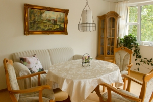 Zimmer mit rundem Tisch, weißer Tischdecke, drei gepolsterte Holzstühle, Sofa mit floralem Kissen, Gemälde an der Wand