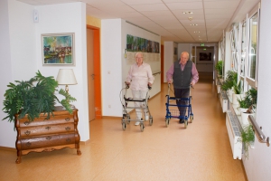 Zwei ältere Menschen gehen mit Rollatoren den hellen Flur entlang, links eine Kommode mit Pflanze und Lampe, rechts Fenster mit weiteren Pflanzen
