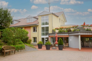 Mehrstöckiges gelbes Gebäude mit großen Fenstern, Terrasse mit Pflanzen und Sitzgelegenheiten, umgeben von Grün