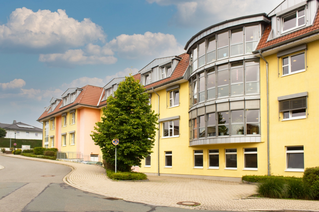 Gelbes Wohngebäude mit großen Glasfenstern und roten Dachziegeln an einer ruhigen Straße unter bewölktem Himmel. Es zeigt die Außenansicht des Haus am Bredberg von der Straße Am Bredberg aus.