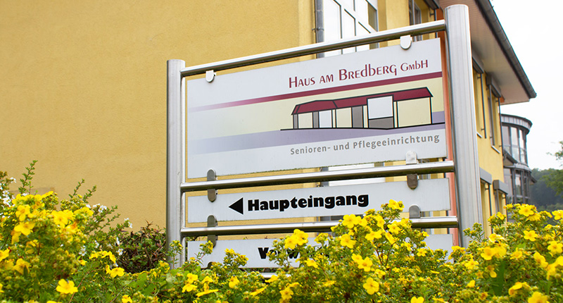 Wegweiser vor gelber Hauswand und gelben Blumen davor, weist zum Haupteingang