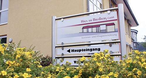 Wegweiser vor gelber Hauswand und gelben Blumen davor, weist zum Haupteingang